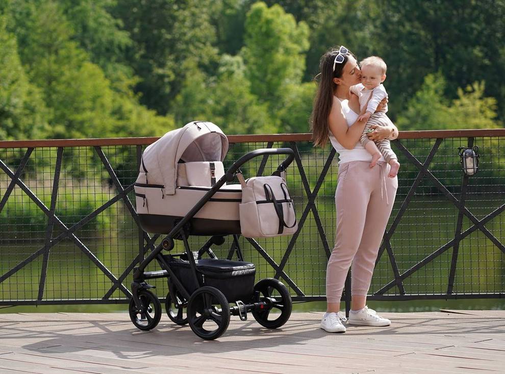 Na zdjęciu różowy wózek 3w1 Camarelo Belagio z lekką gondolą i aluminiowym stelażem, ustawiony nad wodą w spokojnym, zielonym plenerze. Idealny dla dziewczynki.